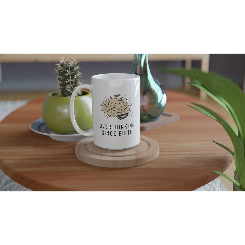 White mug with 'Overthinking Since Birth' text on a wooden table with plants.