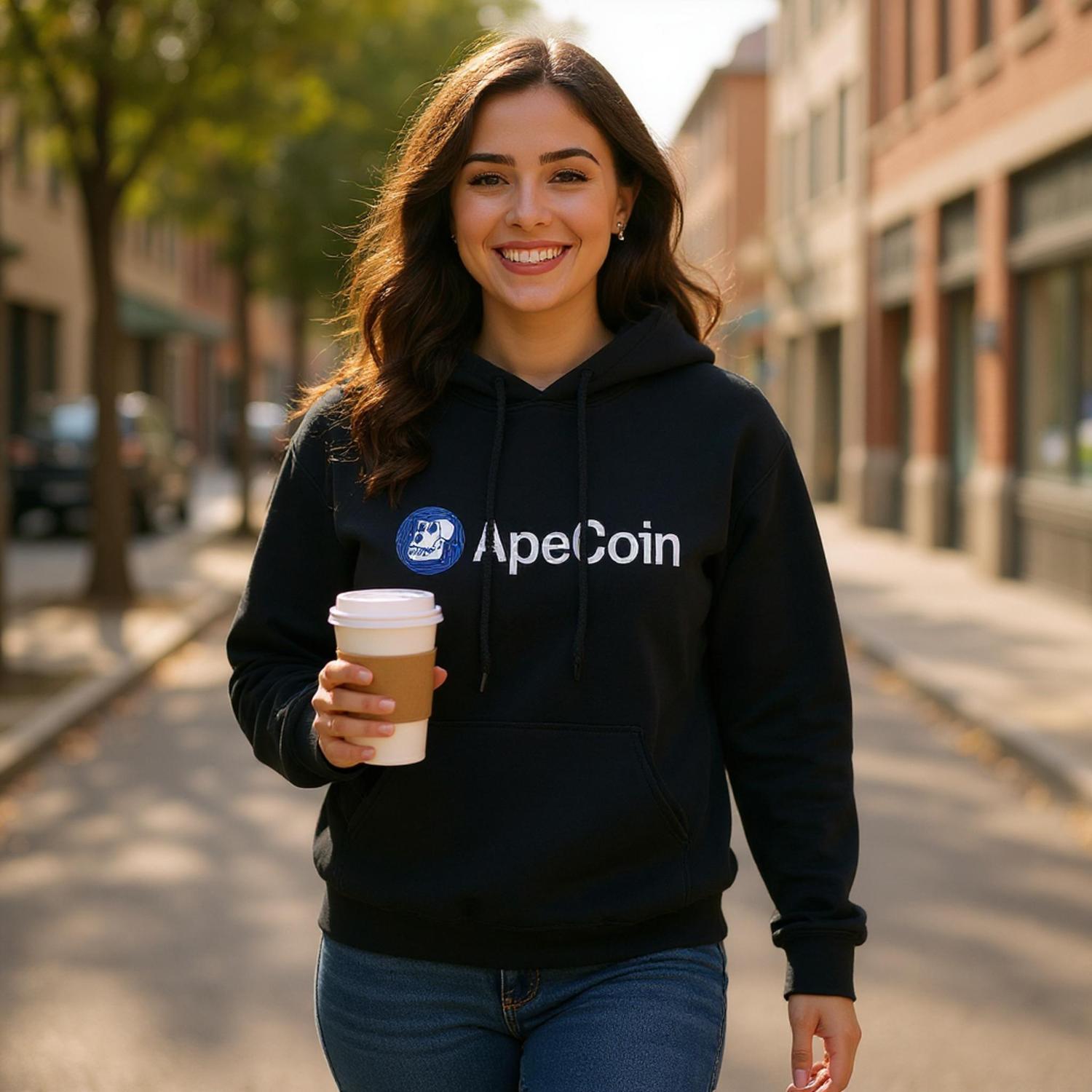 Woman wearing a black hoodie with ApeCoin logo, holding a coffee cup on a city street.