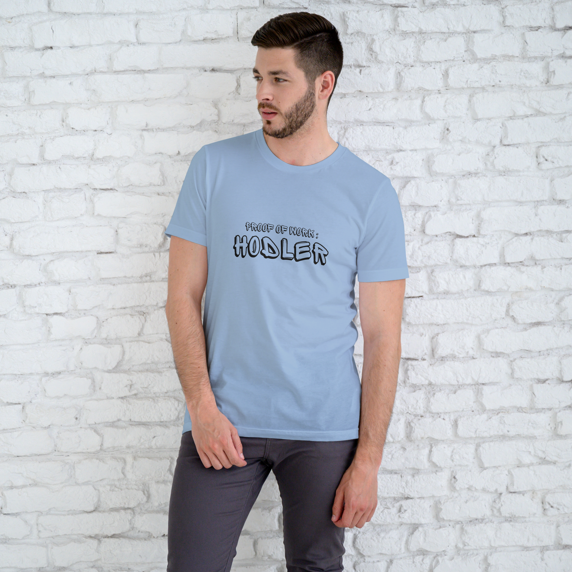 Man wearing a light blue t-shirt with text standing against a white brick wall.