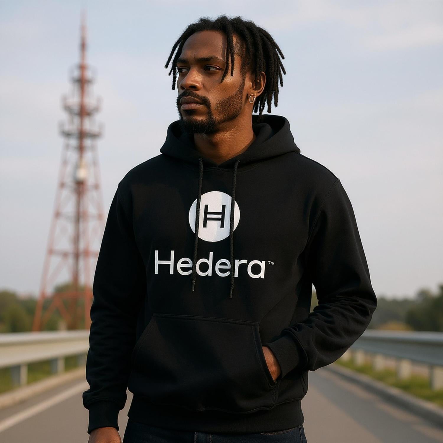 Person wearing a black hoodie with Hedera logo on a road with a tower in the background