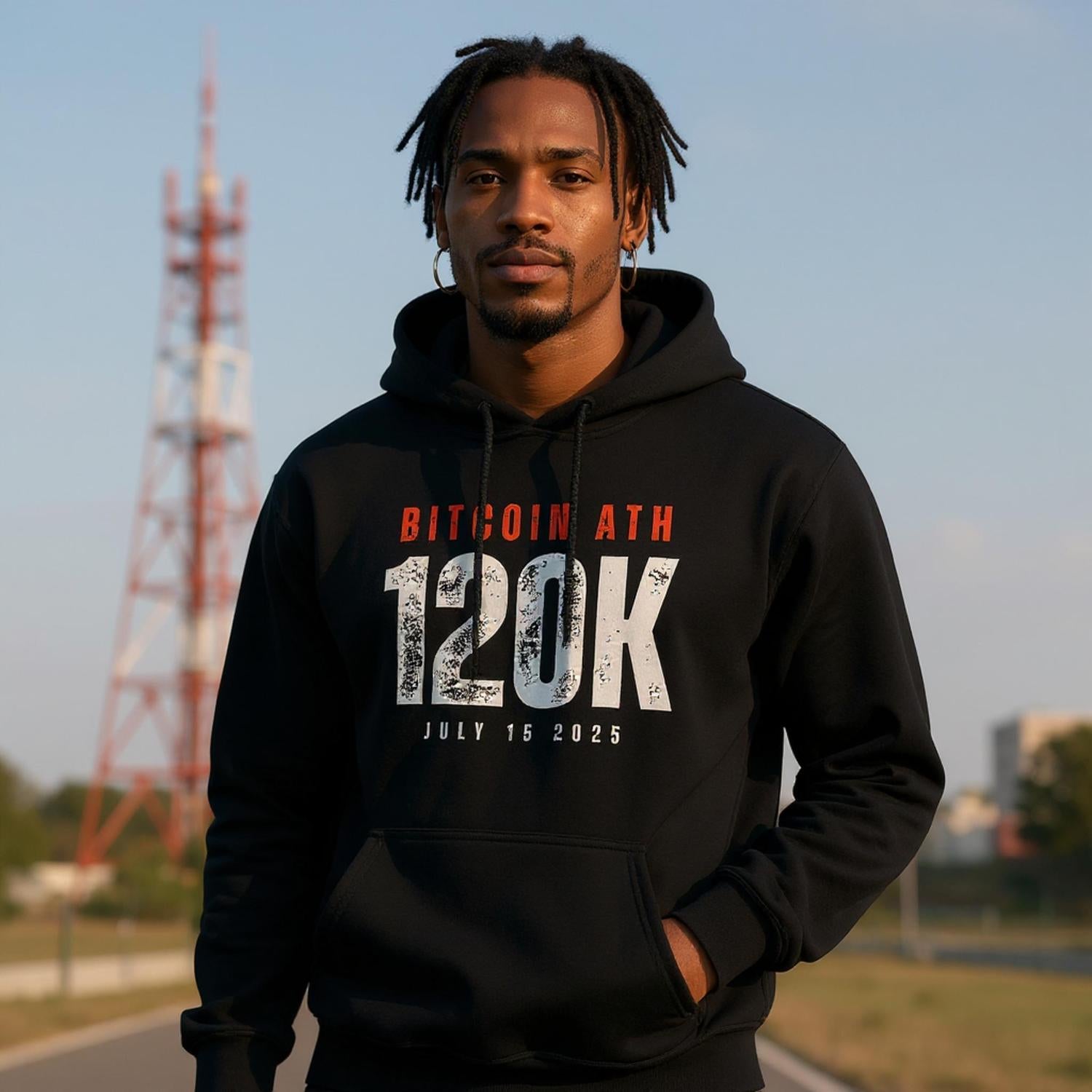 Person wearing a black hoodie with Bitcoin ATH 120K text, standing outdoors with a tower in the background.