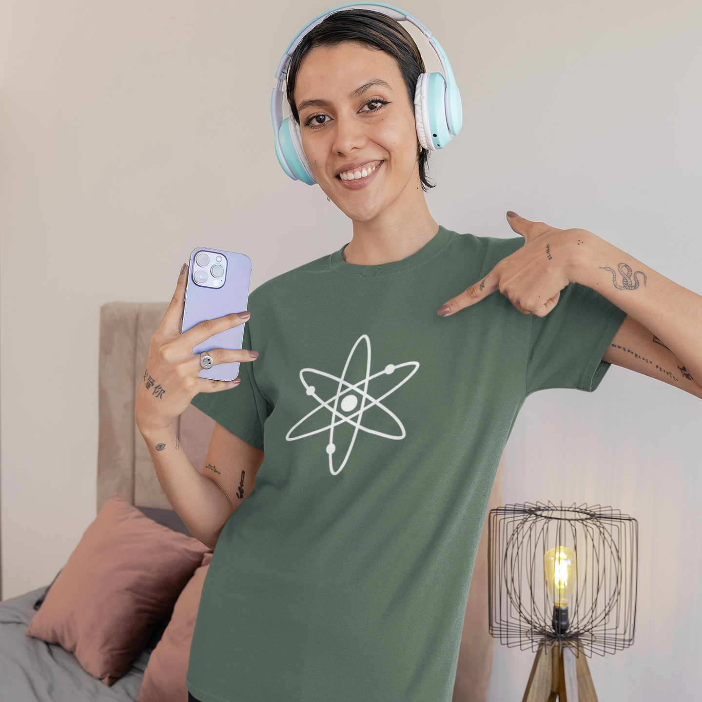 Person wearing a green t-shirt with a white atom symbol, holding a phone in a bedroom setting.
