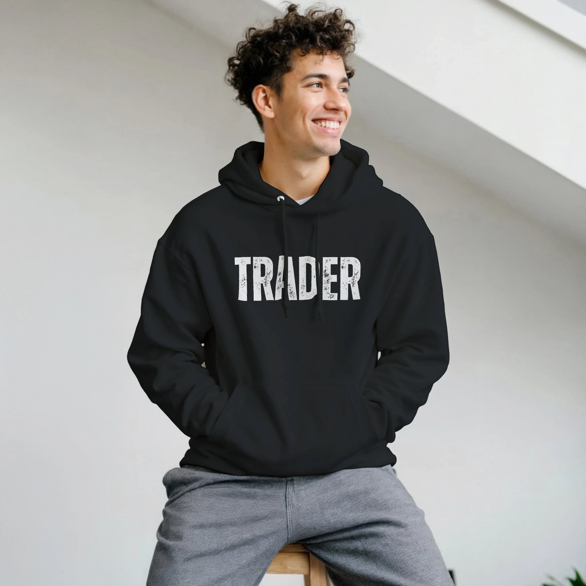 Man wearing a black hoodie with 'TRADER' text, sitting on a white surface.