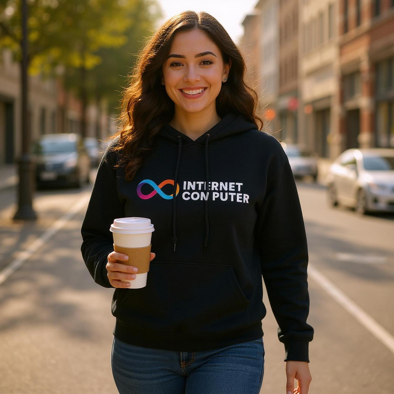 Woman wearing a black hoodie with 'INTERNET COMPUTER' text and logo, holding a coffee cup on a city street.