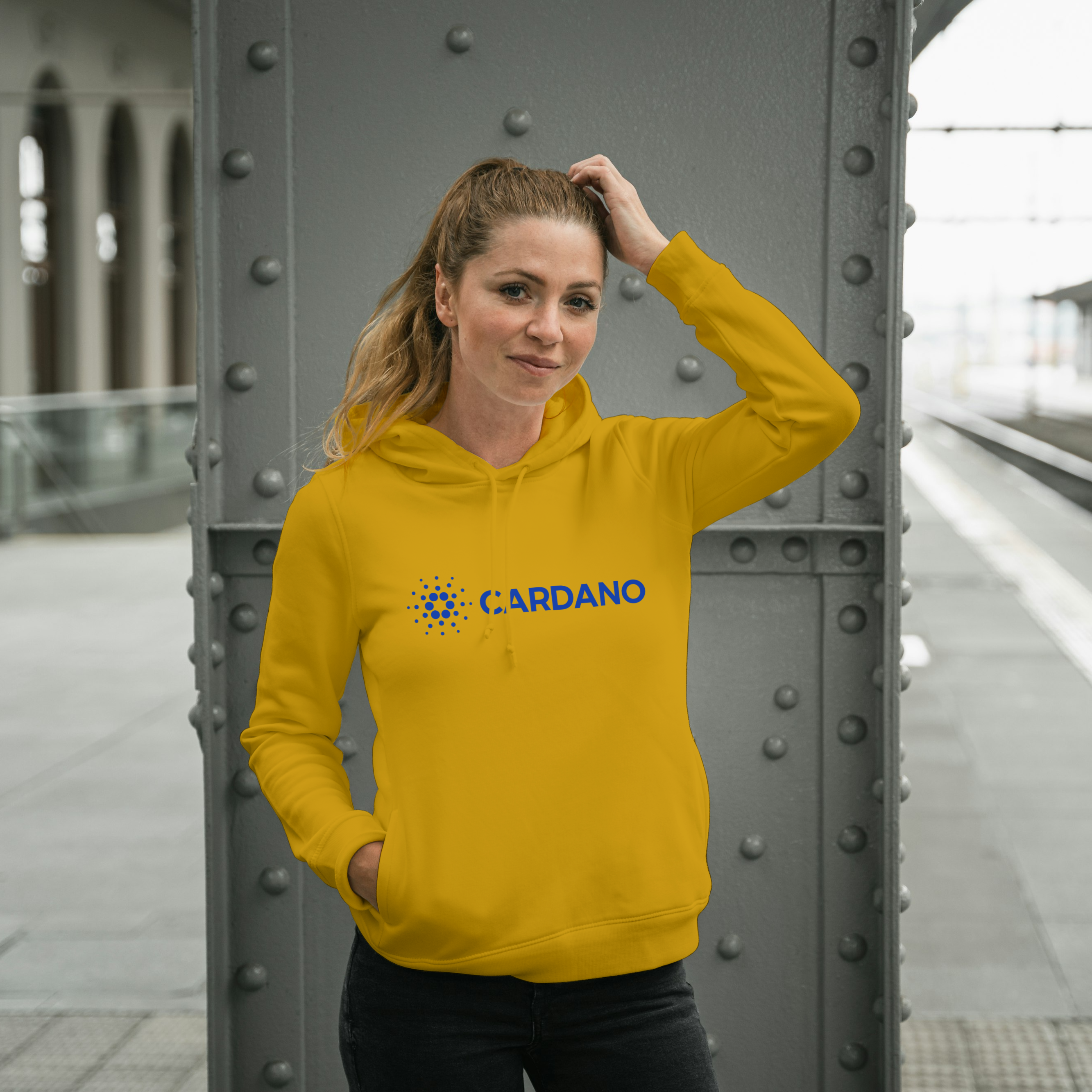 Person wearing a yellow hoodie with 'Cardano' logo at a train station.