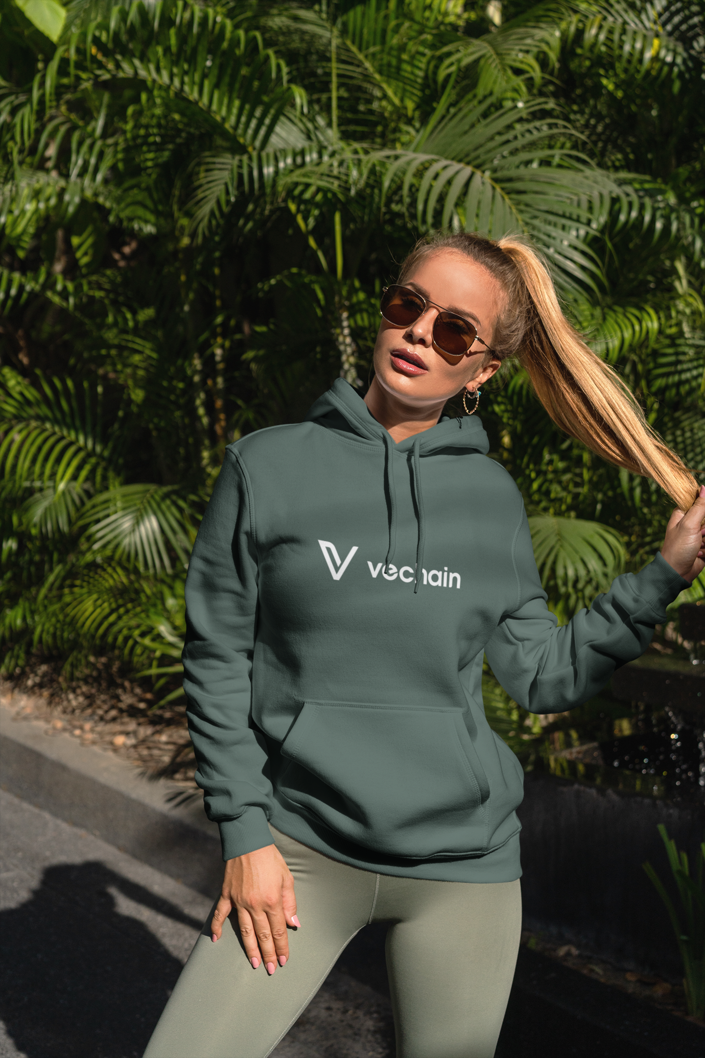 Woman wearing a green hoodie with a logo, standing outdoors with greenery in the background