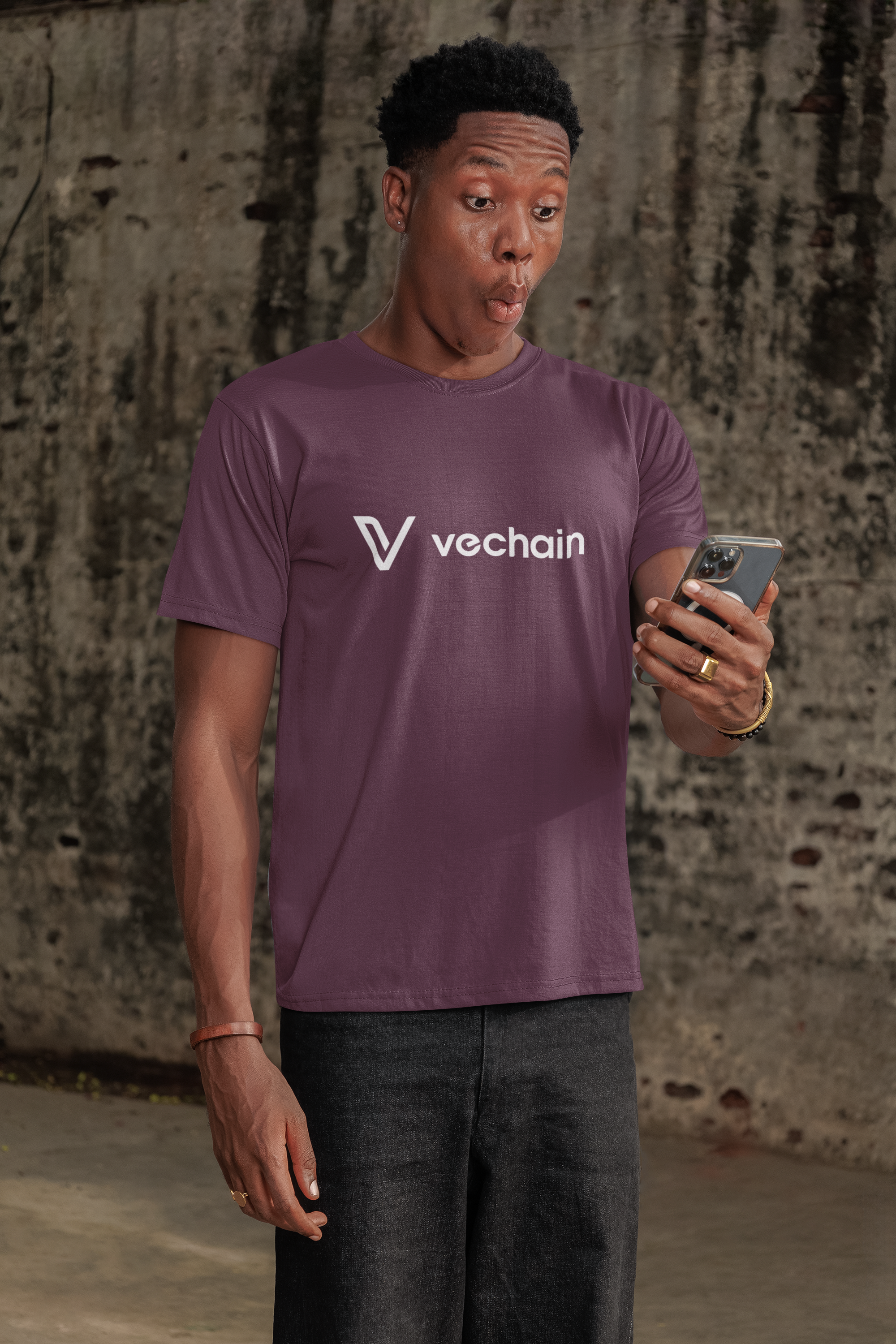 Person wearing maroon t-shirt with 'vechain' logo, holding a phone against a textured wall.