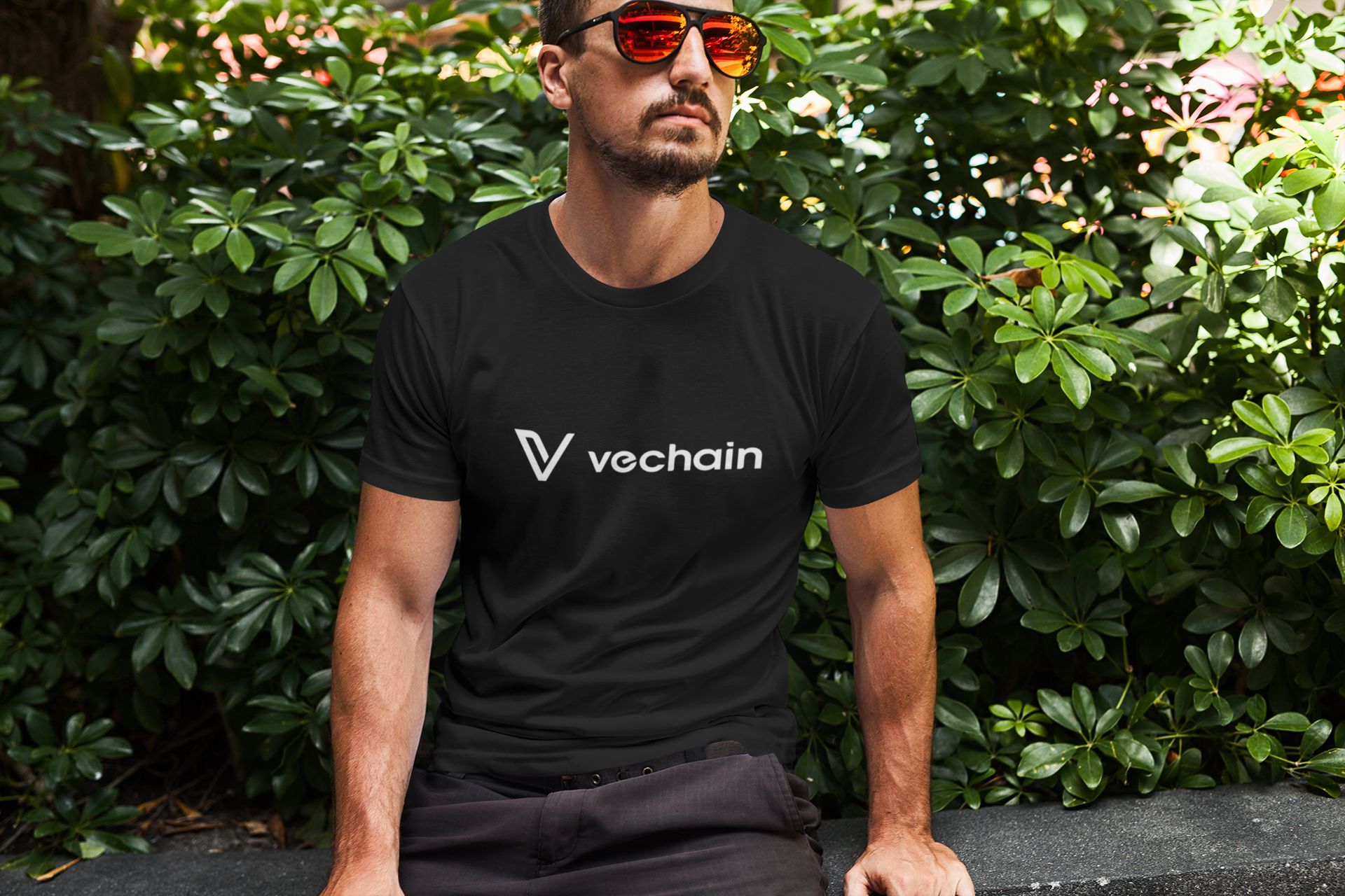 Man wearing a black t-shirt with 'vechain' logo sitting outdoors among green bushes.