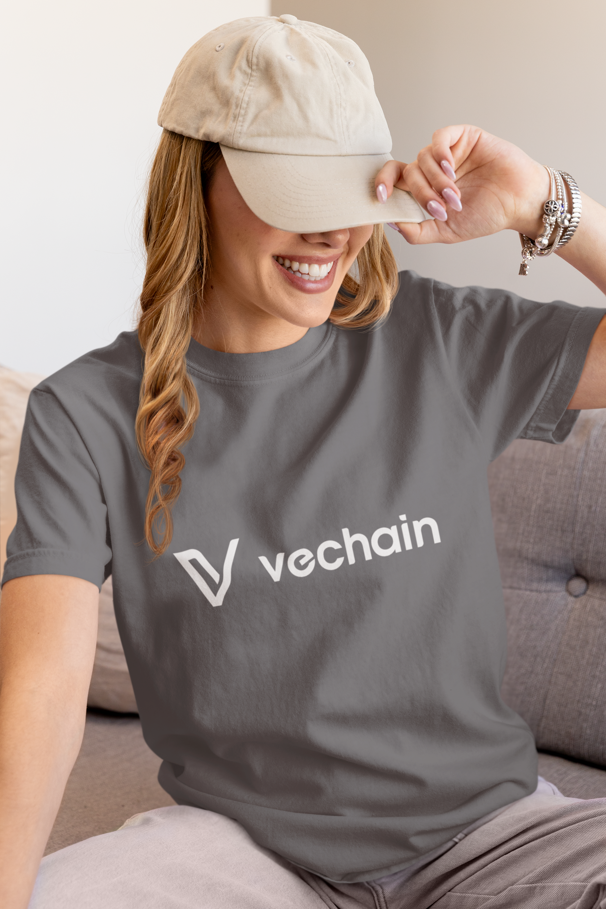 Person wearing a gray t-shirt with 'vechain' logo and beige cap, sitting on a couch.