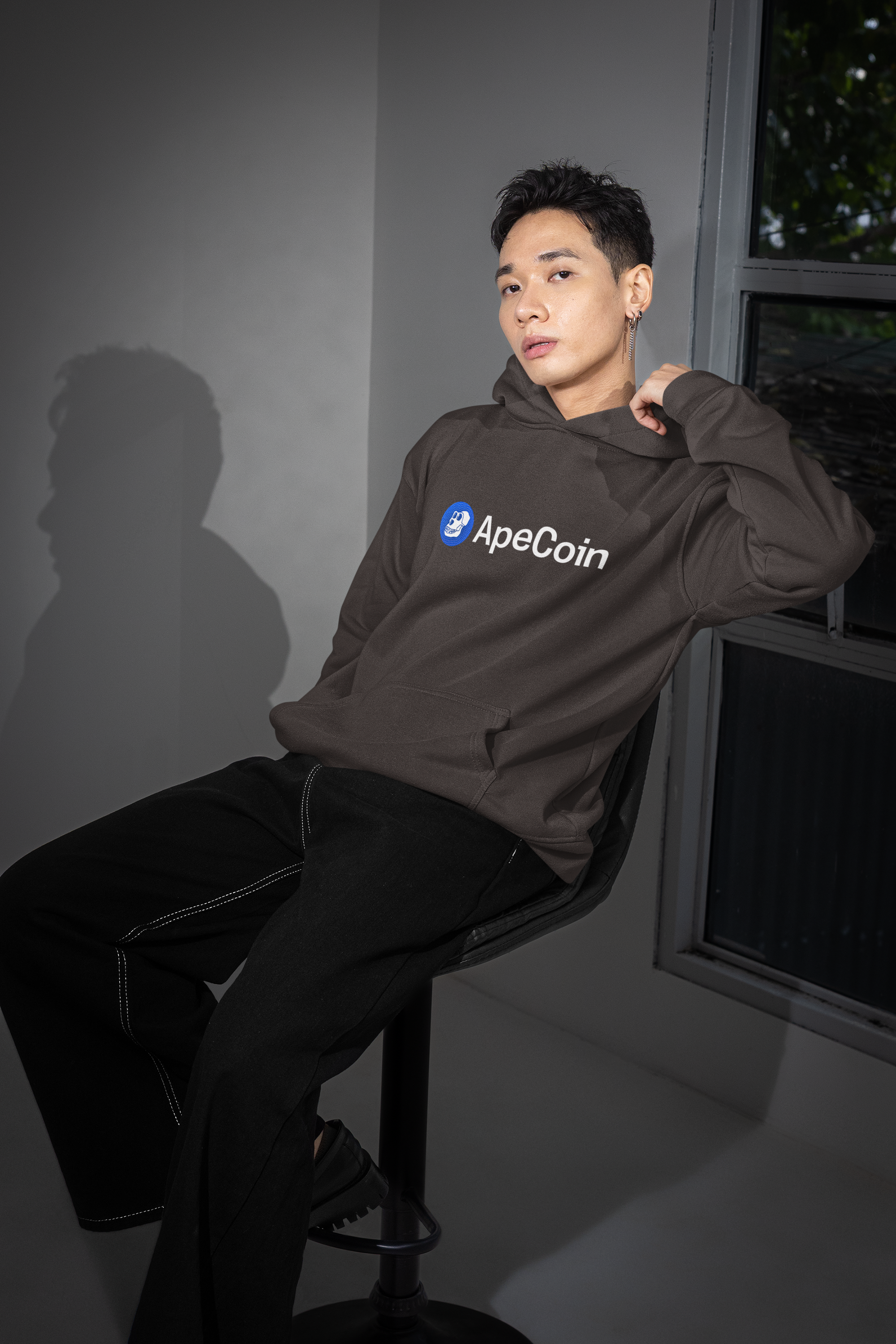 Person wearing a hoodie with ApeCoin logo sitting on a stool against a dark background