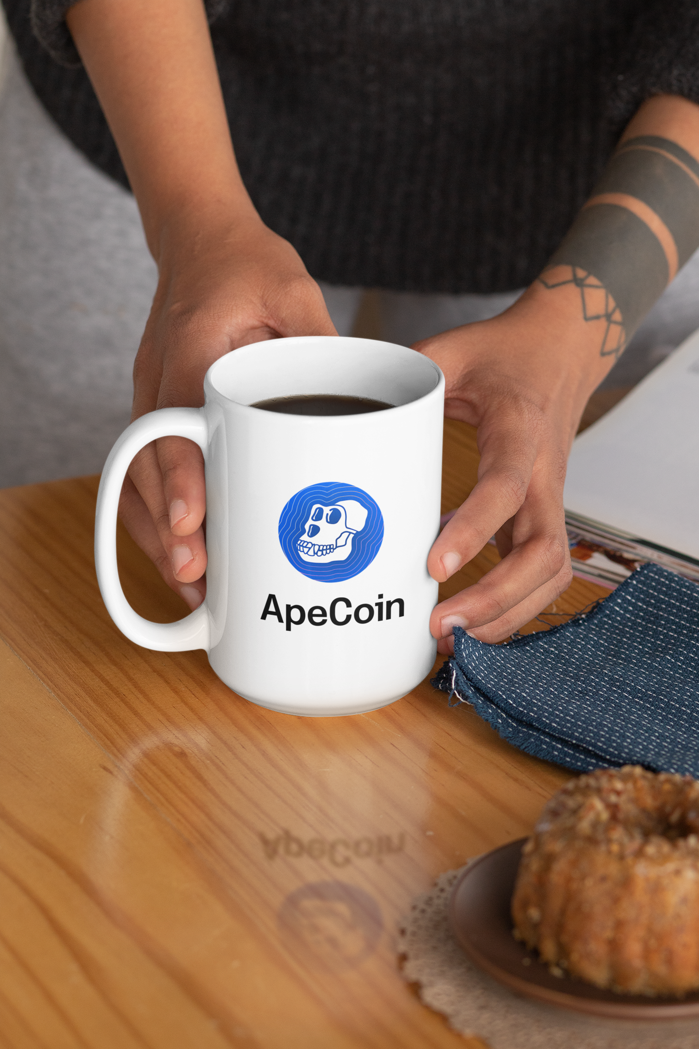 Person holding a white mug with ApeCoin logo on a wooden table.