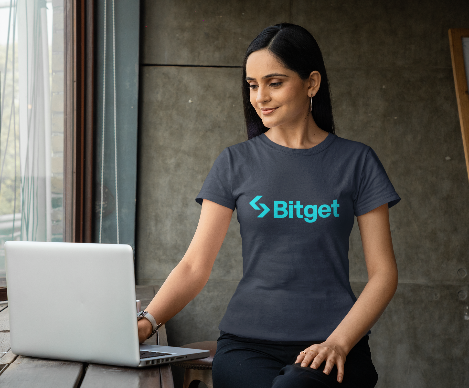 Woman wearing a dark blue t-shirt with Bitget logo using a laptop.
