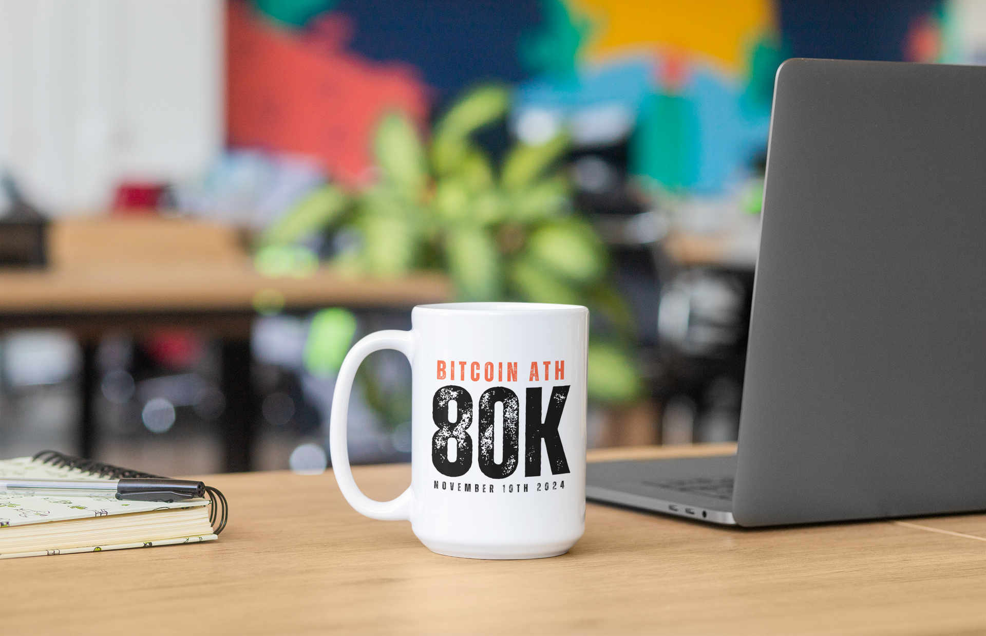 Mug with Bitcoin AT 80K text on a desk with a laptop and notebook
