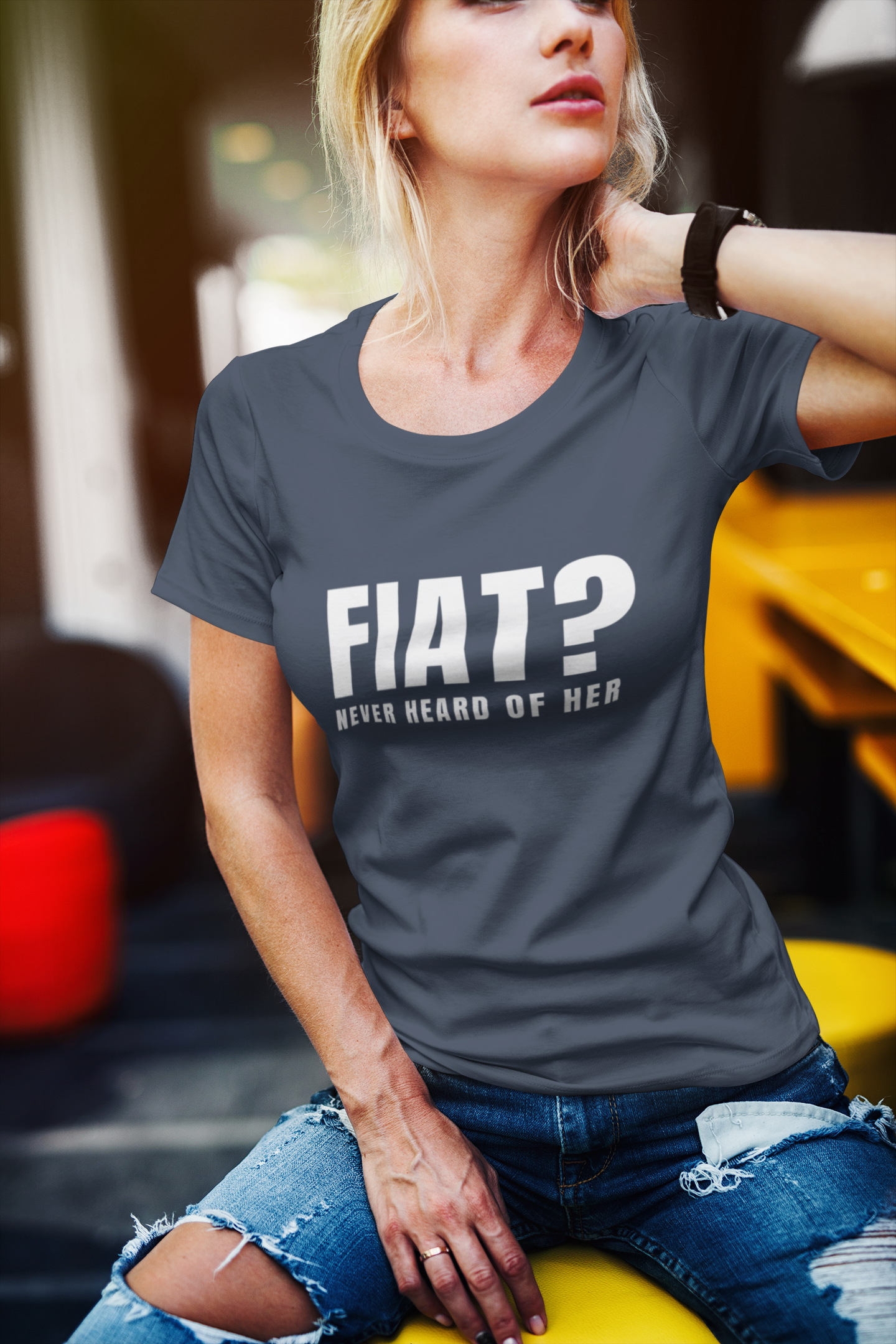 Woman wearing a gray t-shirt with 'FIAT? Never Heard of Her' text, sitting in front of a yellow car.