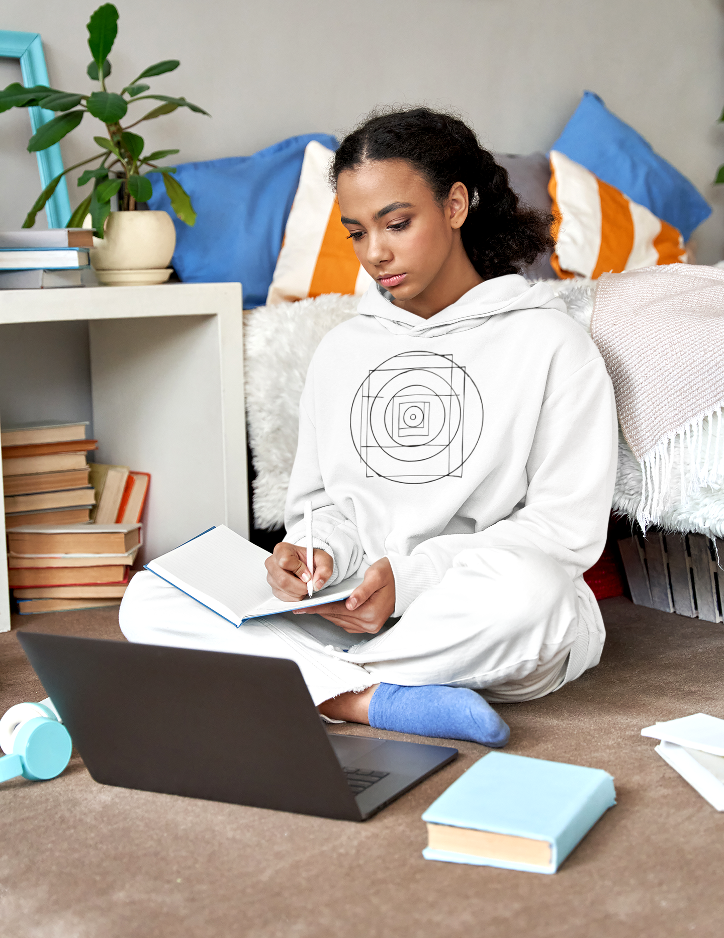 Woman in a white hoodie sitting on the floor with a laptop and books, surrounded by colorful pillows and a plant.