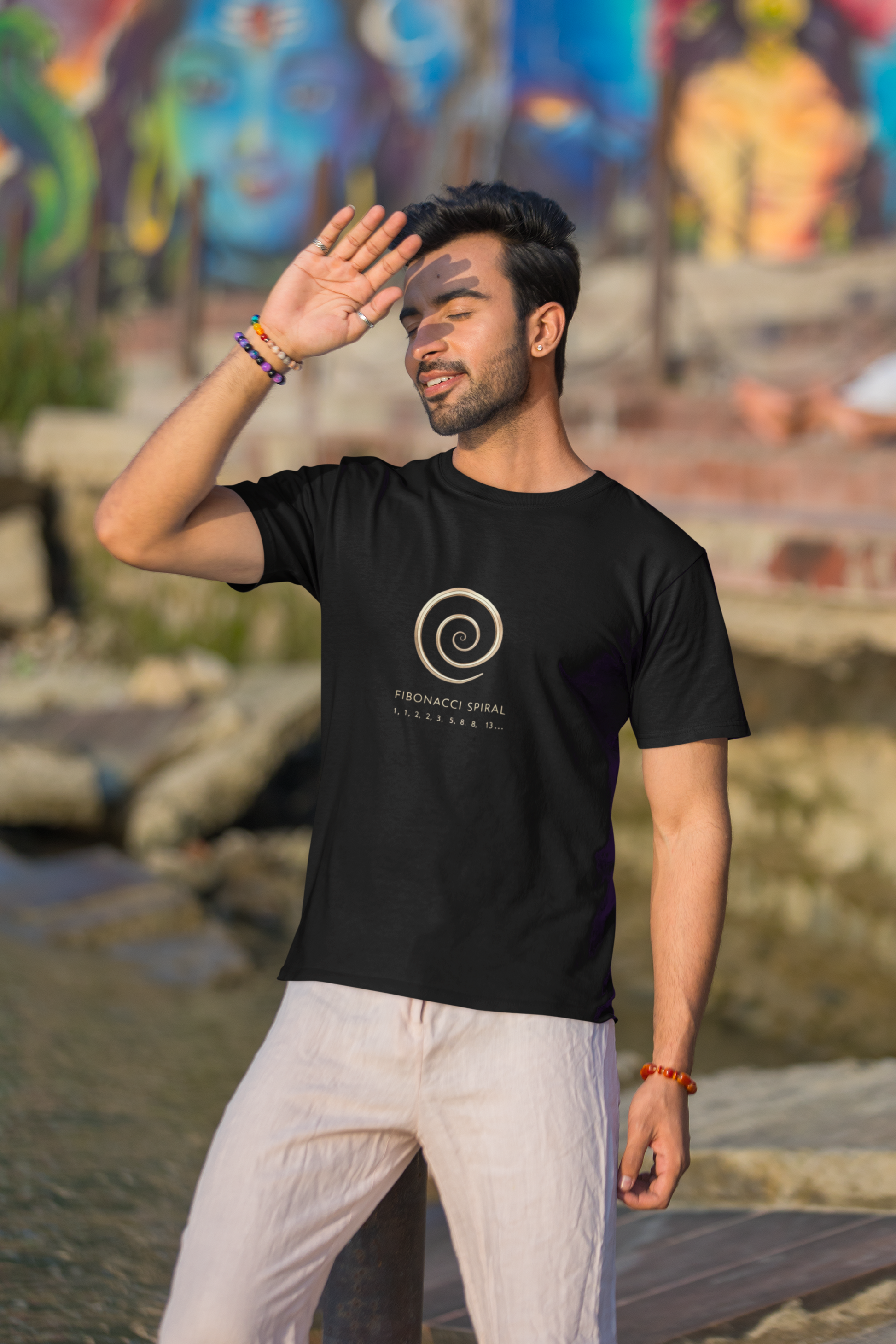Man wearing a black t-shirt with a logo, standing outdoors with a colorful mural in the background.