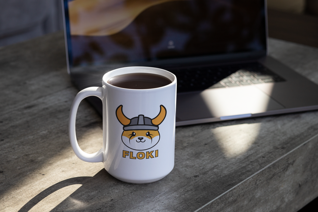 Mug with a cartoon dog wearing a helmet and 'FLOKI' text on a wooden table with a laptop in the background.