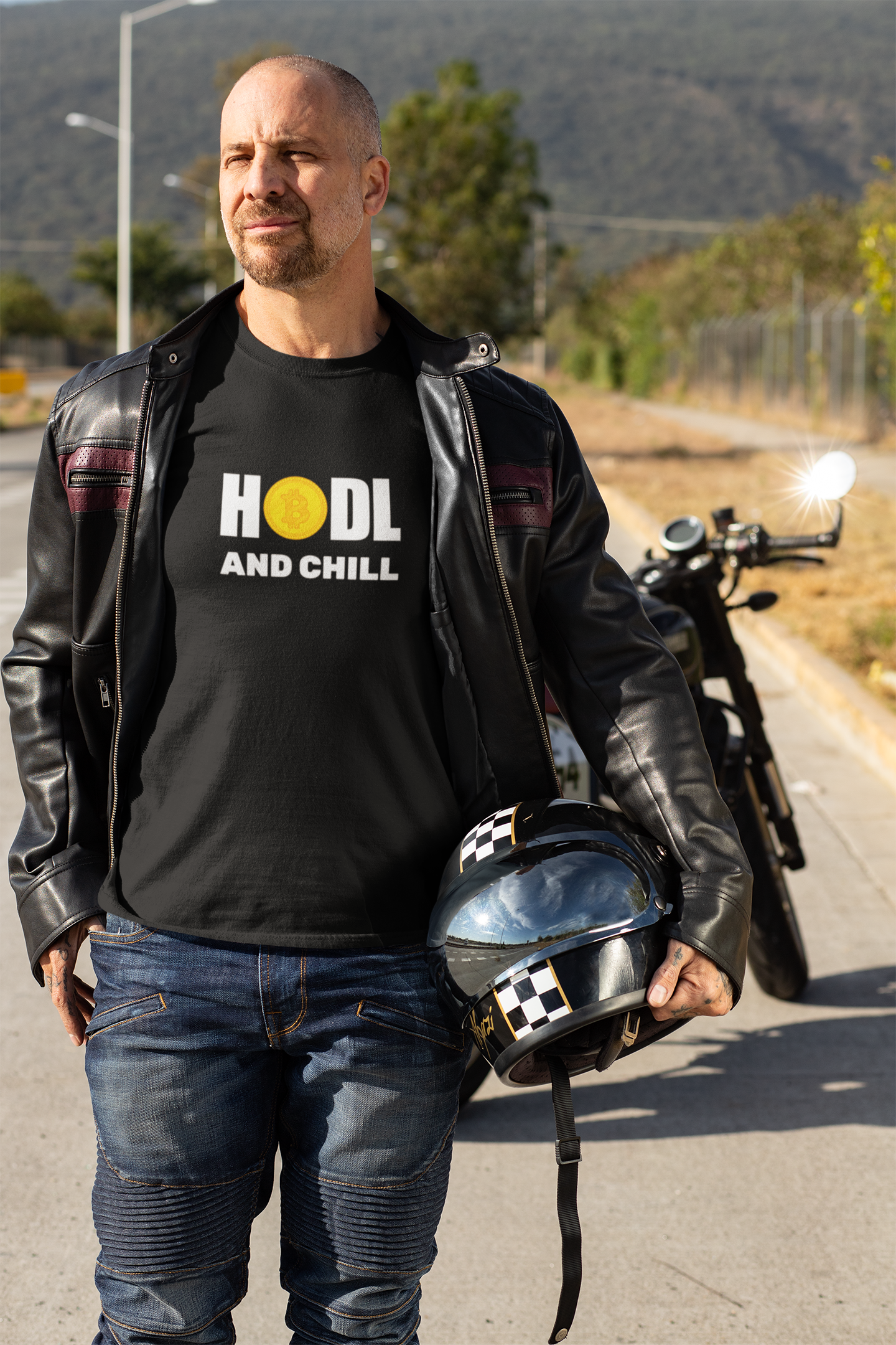 Man wearing a black 'HODL AND CHILL' t-shirt, holding a helmet, with a motorcycle in the background.
