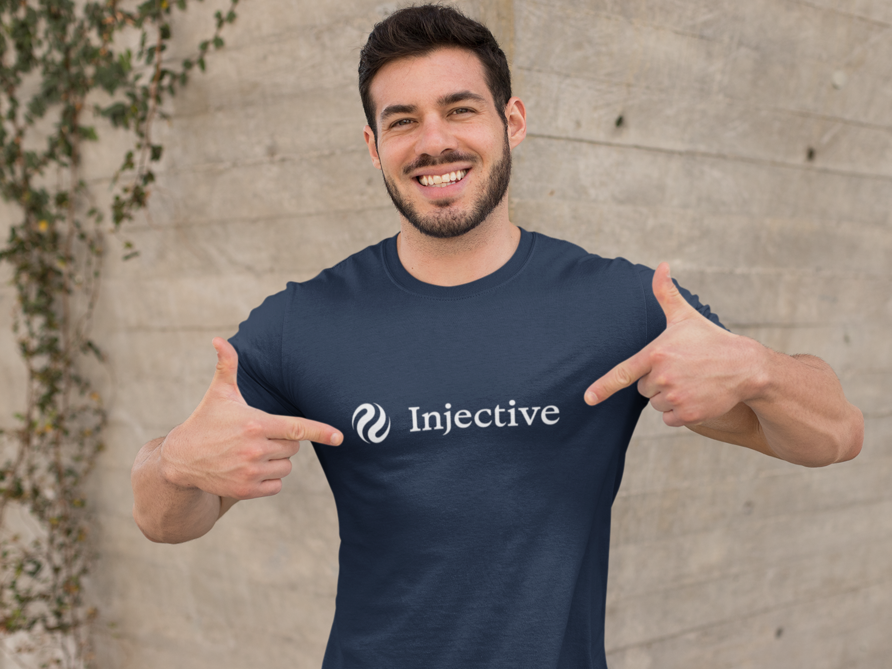 Man wearing a navy blue t-shirt with 'Injective' logo, pointing at it, against a concrete wall.