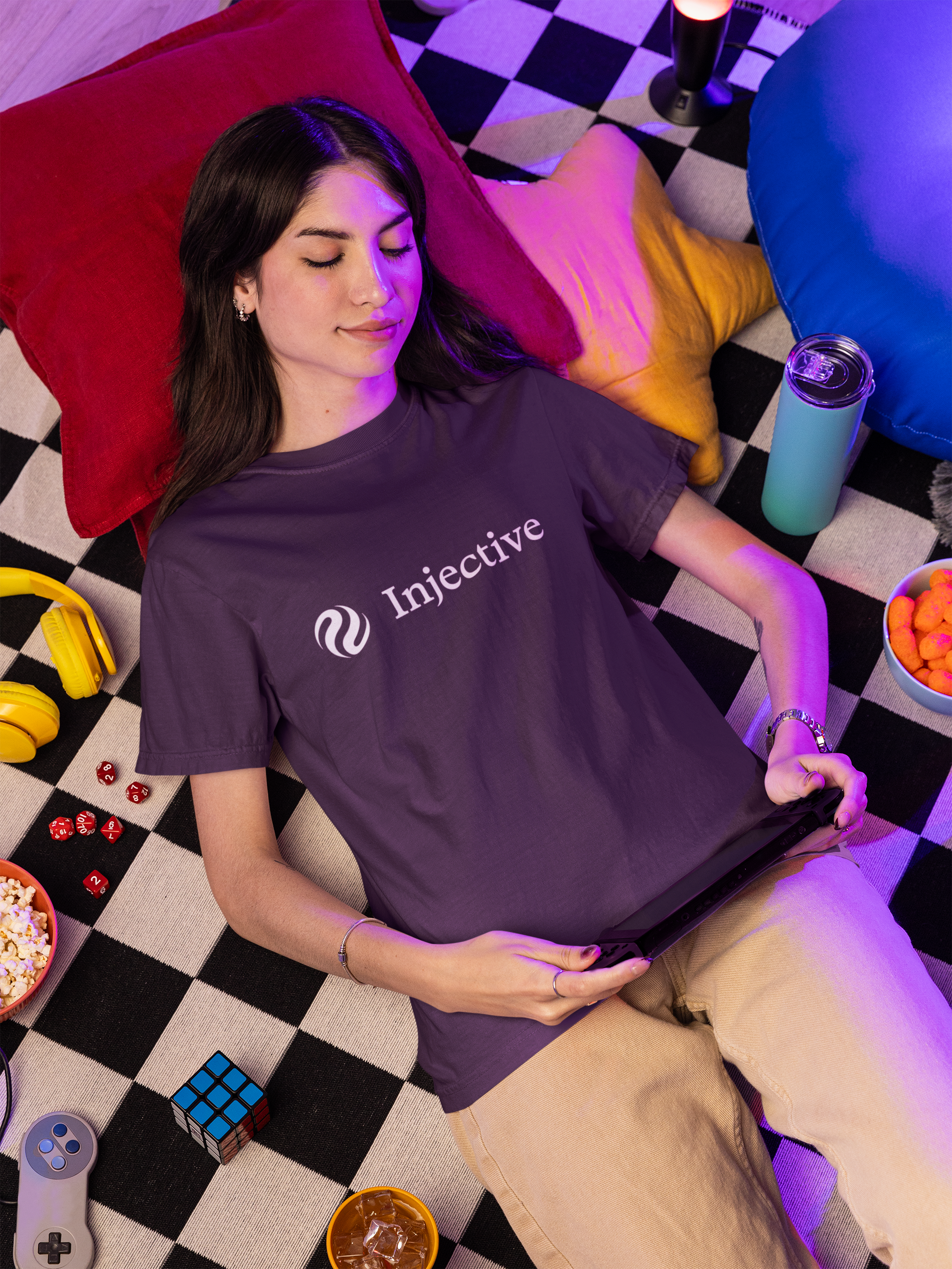 Person wearing a purple 'Injective' t-shirt lying on a checkered floor with colorful pillows and snacks.