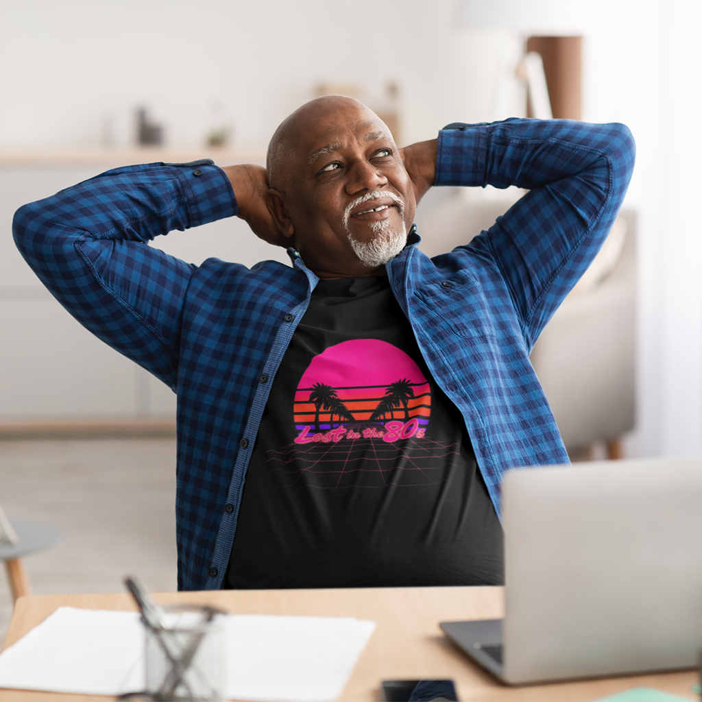 Man sitting at a desk with a laptop, wearing a colorful t-shirt and blue jacket.