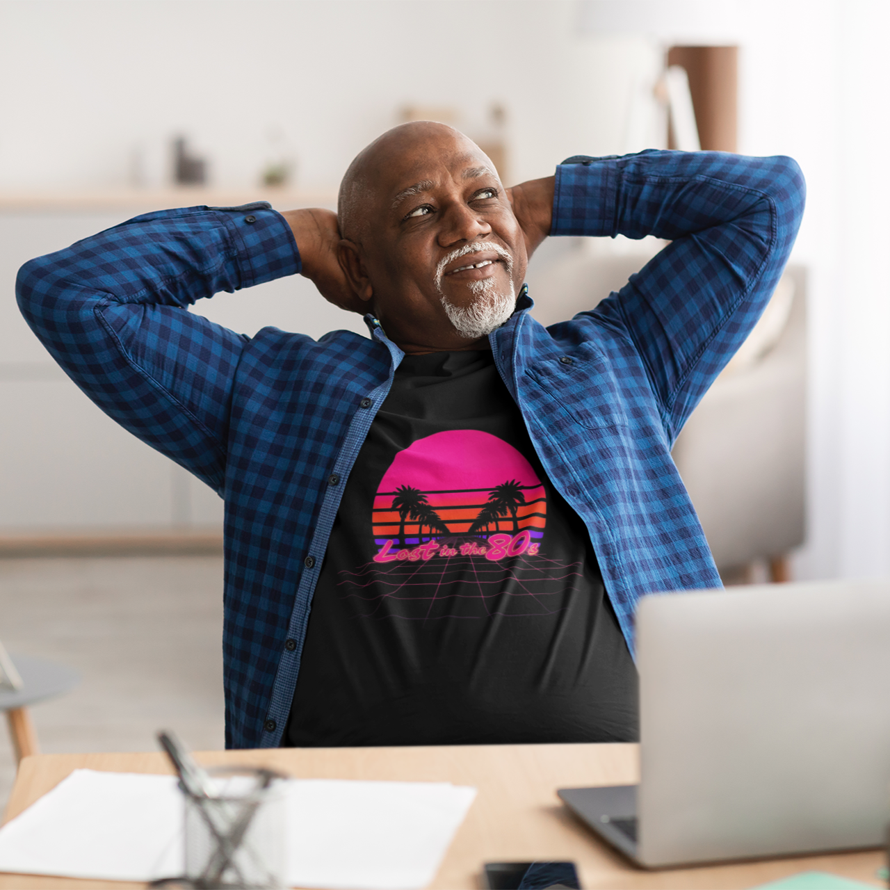 Man sitting at a desk with a laptop, wearing a colorful t-shirt and blue jacket.