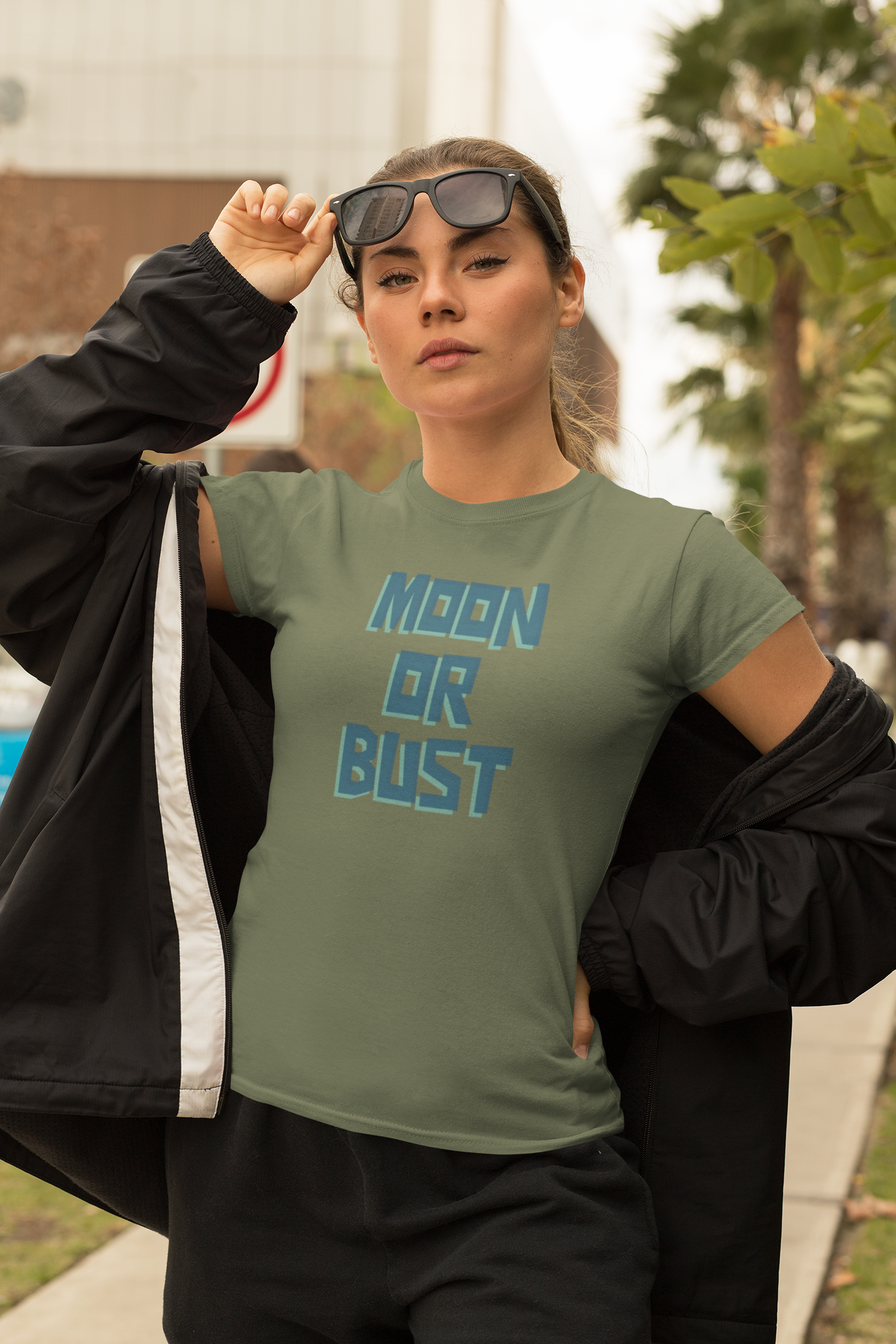 Person wearing a green t-shirt with 'MOON OR BUST' text, holding sunglasses, outdoors.