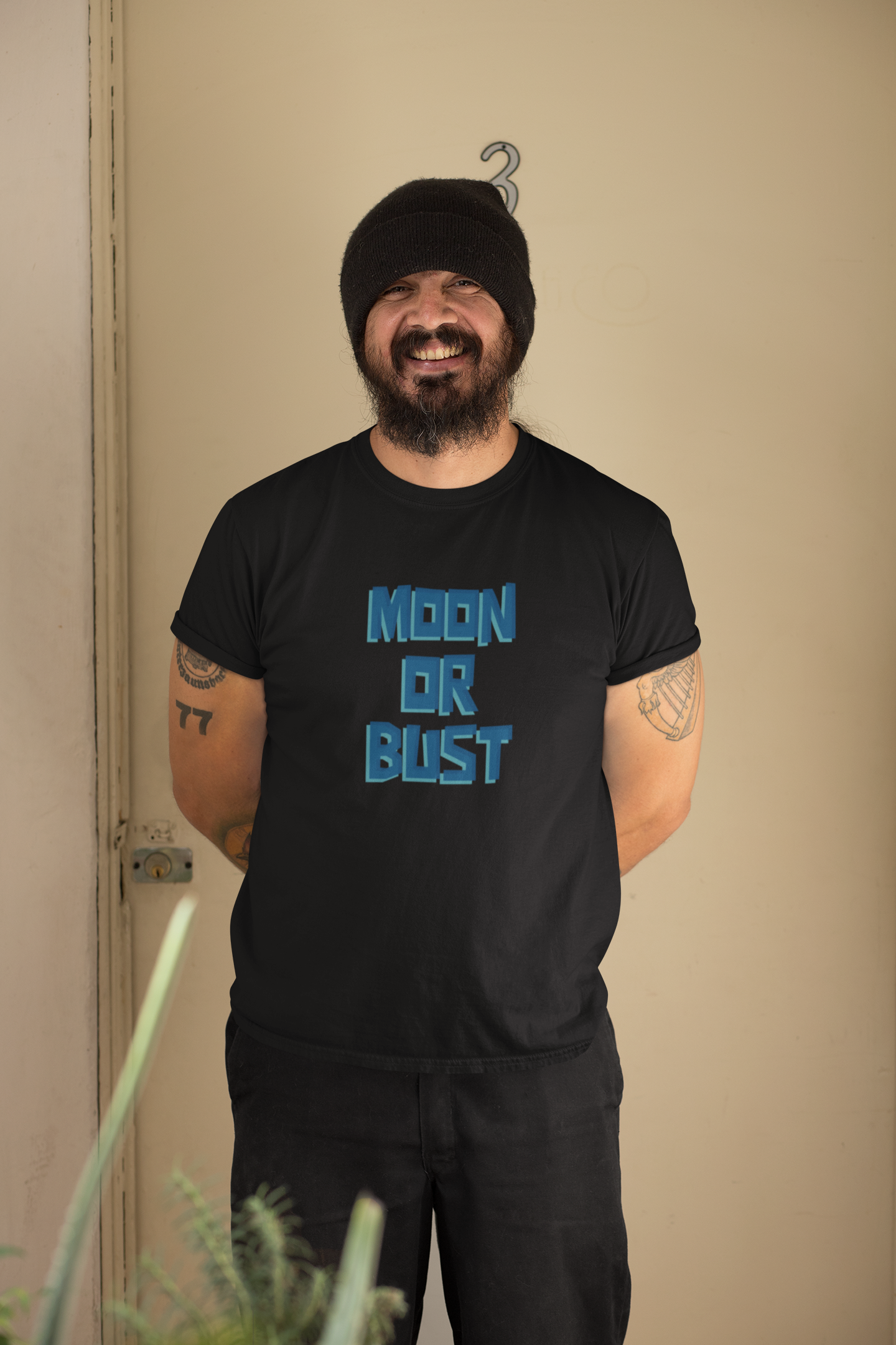 Man wearing a black t-shirt with 'MOON OR BLUST' text, standing against a beige wall.