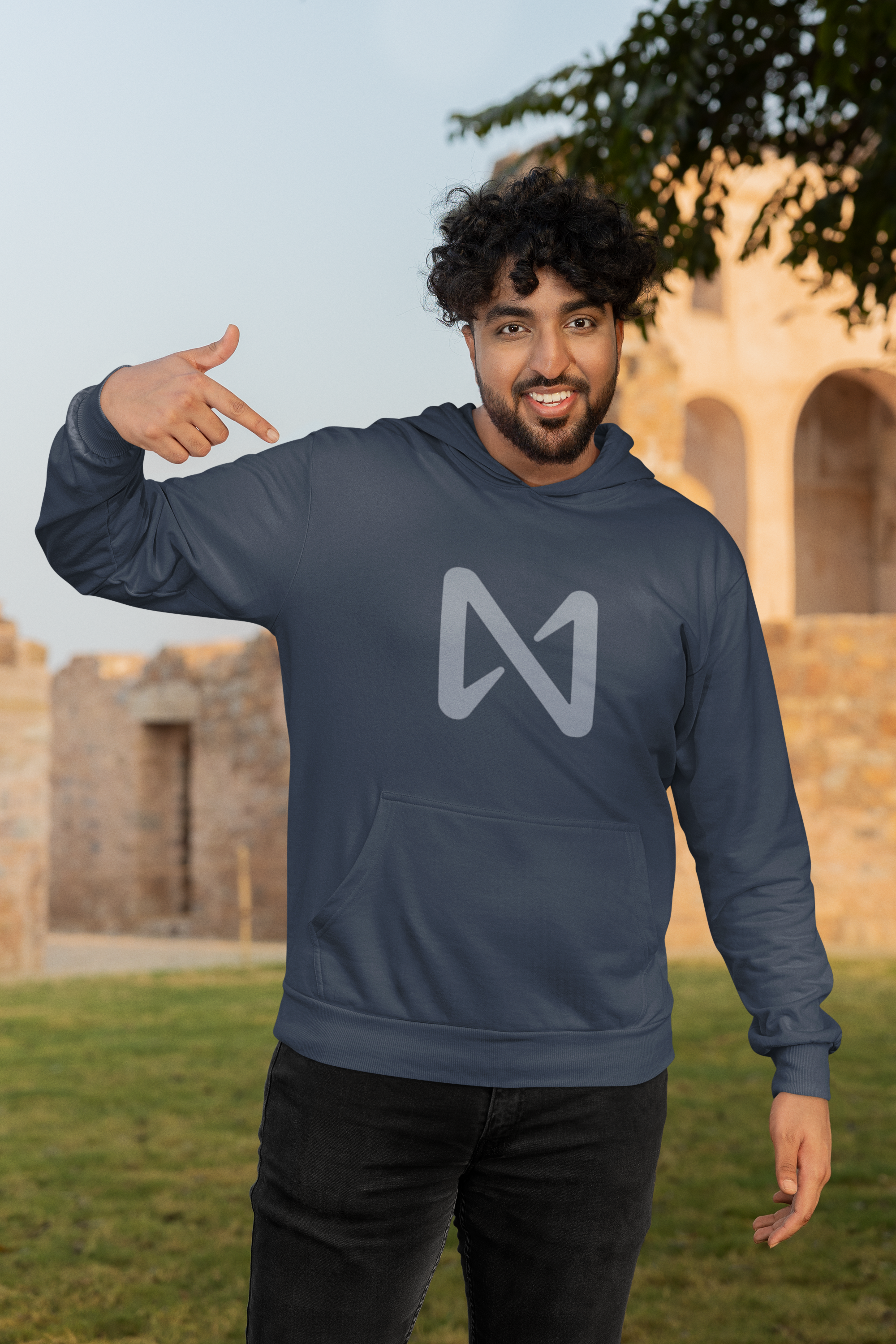 Man wearing a blue hoodie with a logo, pointing to it, in an outdoor setting.