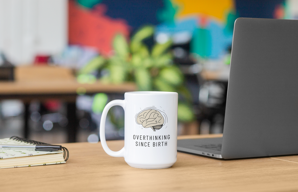 White mug with brain graphic and text 'Overthinking Since Birth' on a desk with a laptop and notebook.