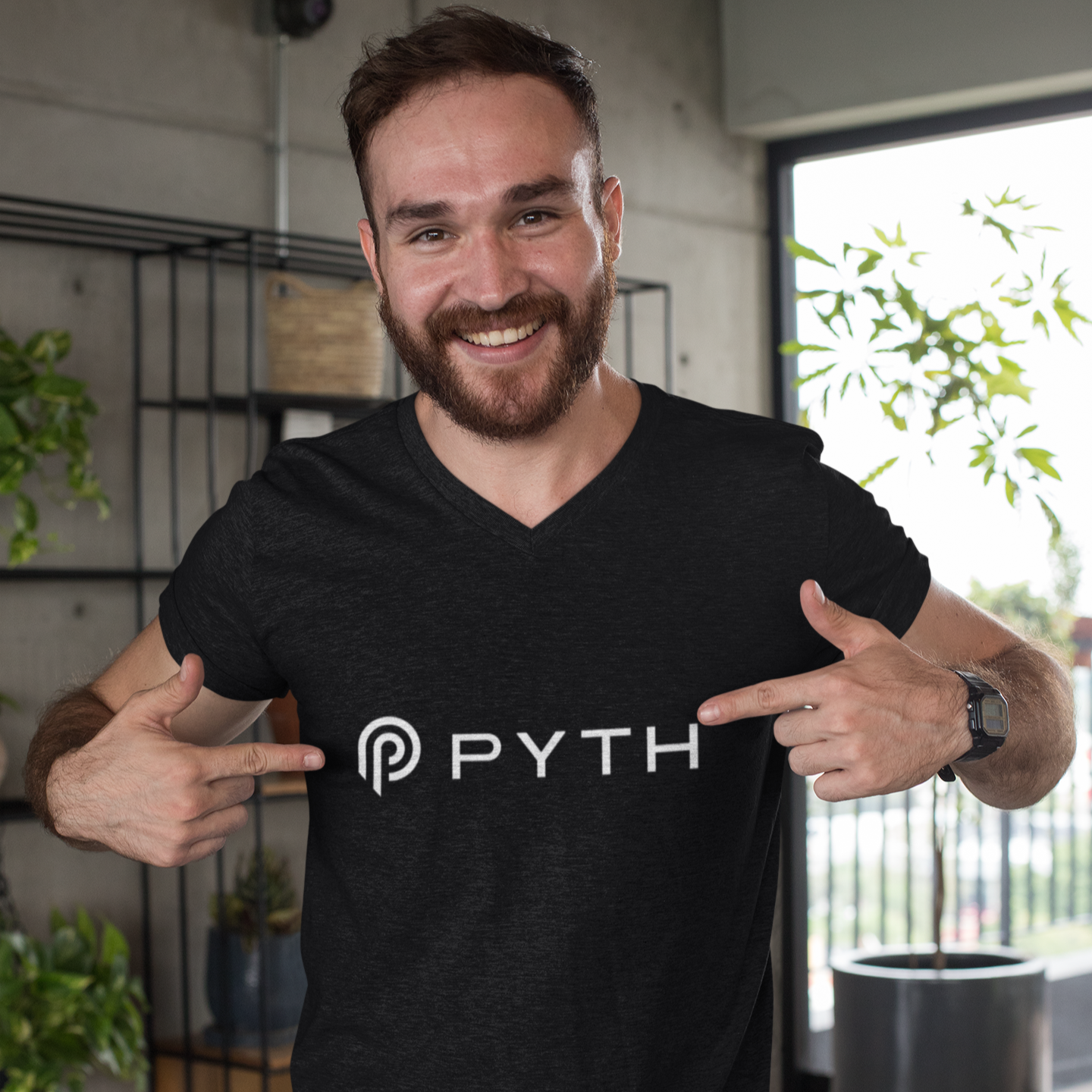 Man wearing a black t-shirt with 'PYTH' logo, pointing at it, in an indoor setting.