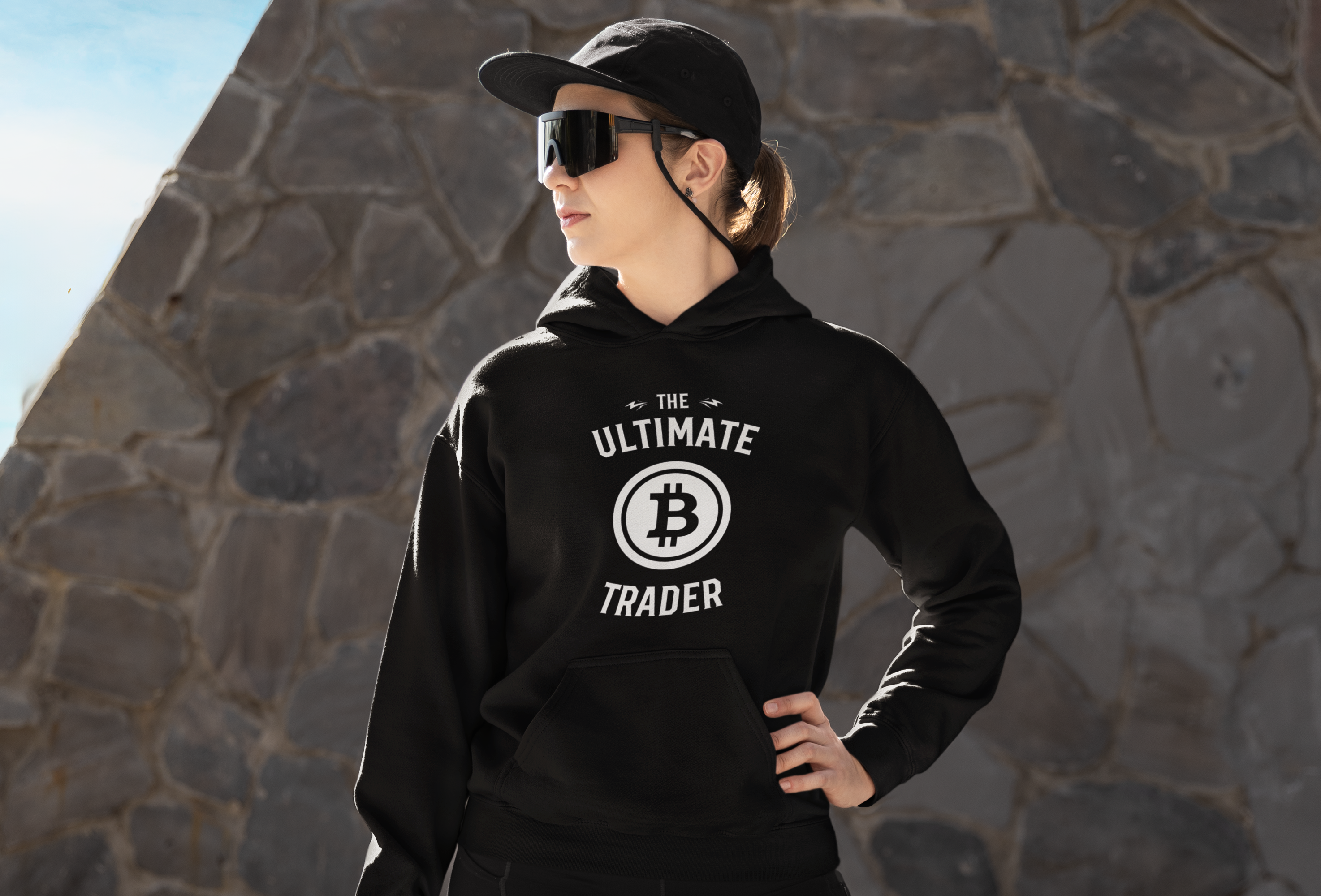 Woman wearing a black hoodie with 'The Ultimate Trader' and Bitcoin logo, standing against a stone wall.