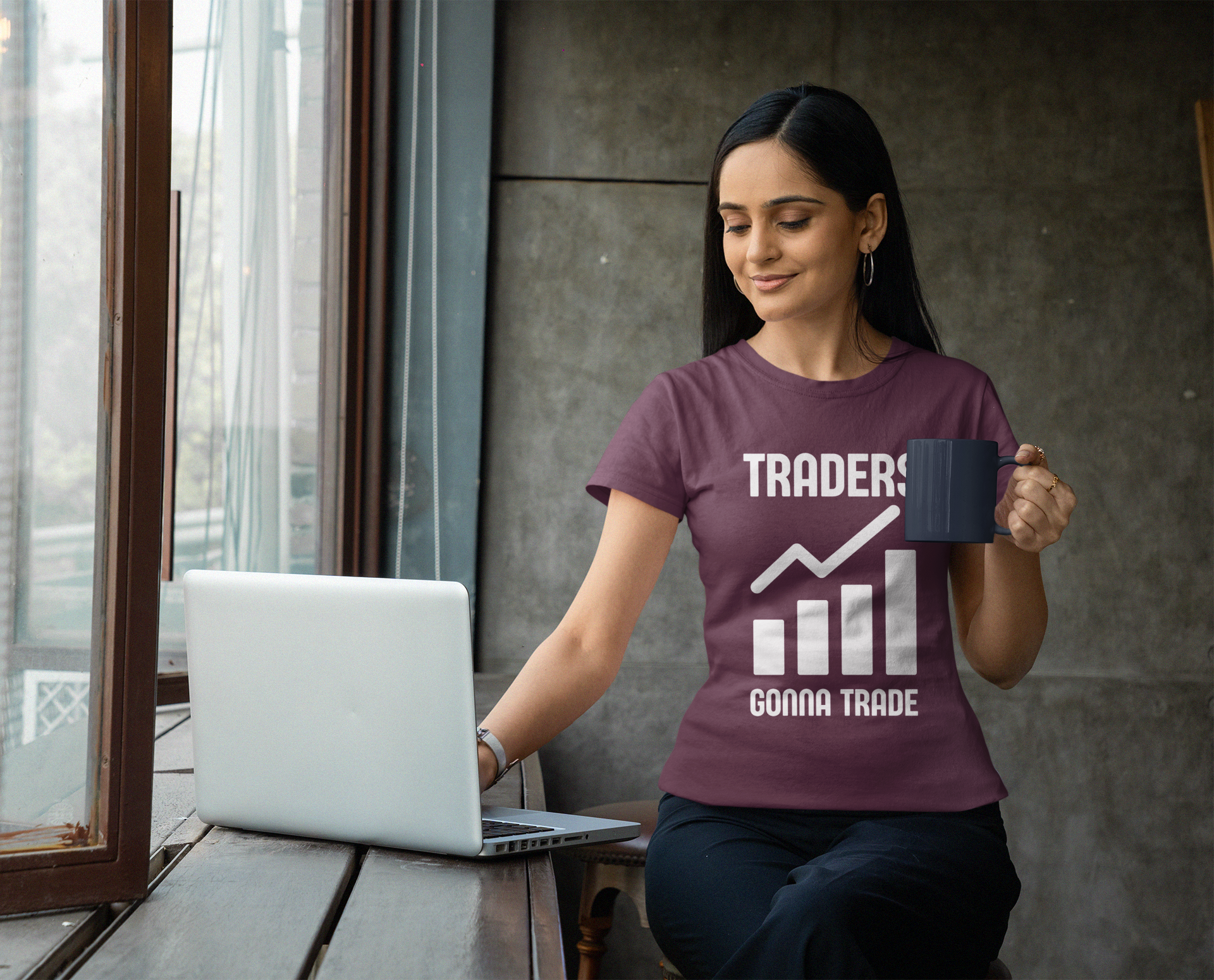 Woman wearing a t-shirt with 'TRADERS GONNA TRADE' holding a mug and using a laptop.
