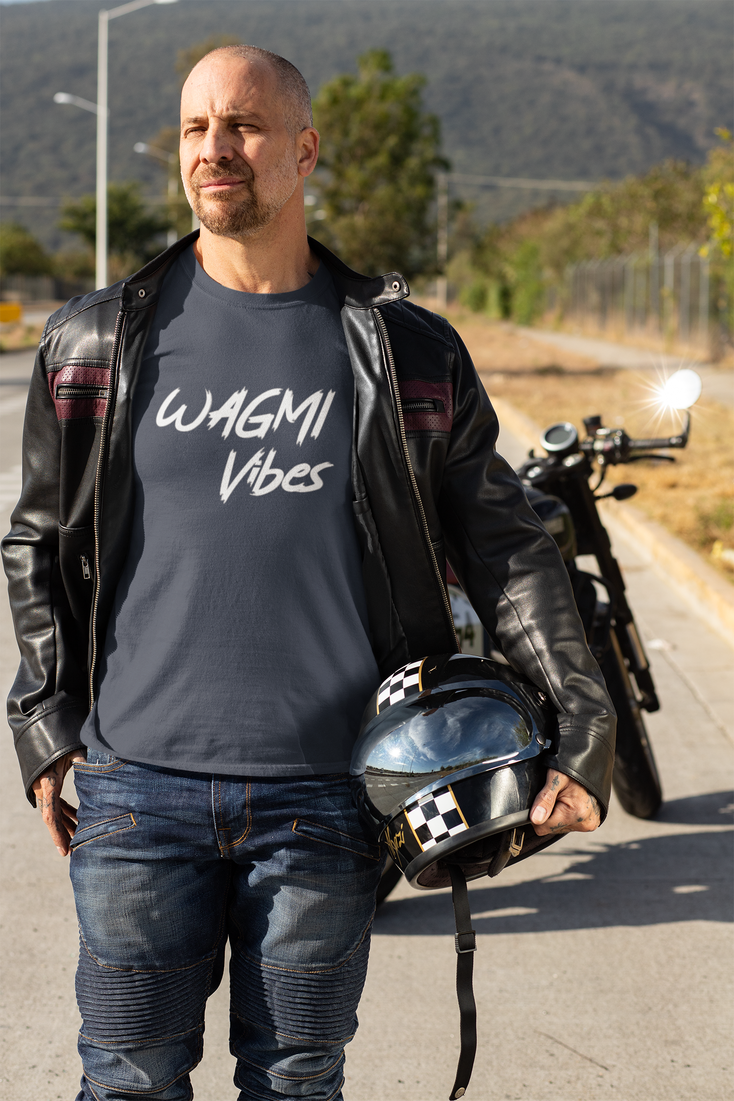 Man in leather jacket and 'WAGMI Vibes' t-shirt holding a helmet on a road with a motorcycle in the background.