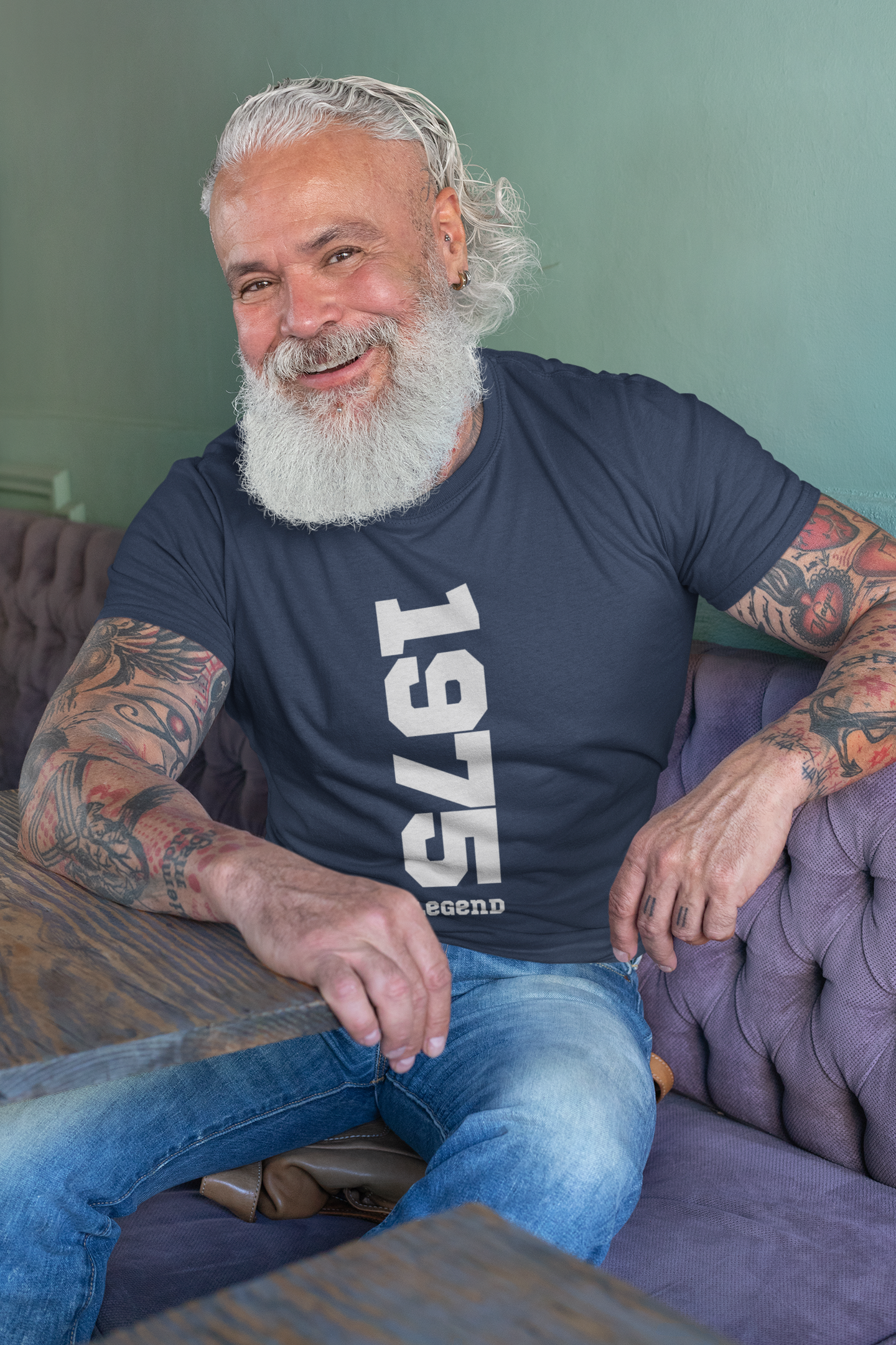 Man with tattoos and a beard wearing a blue t-shirt with '1975' printed on it, sitting on a purple couch.