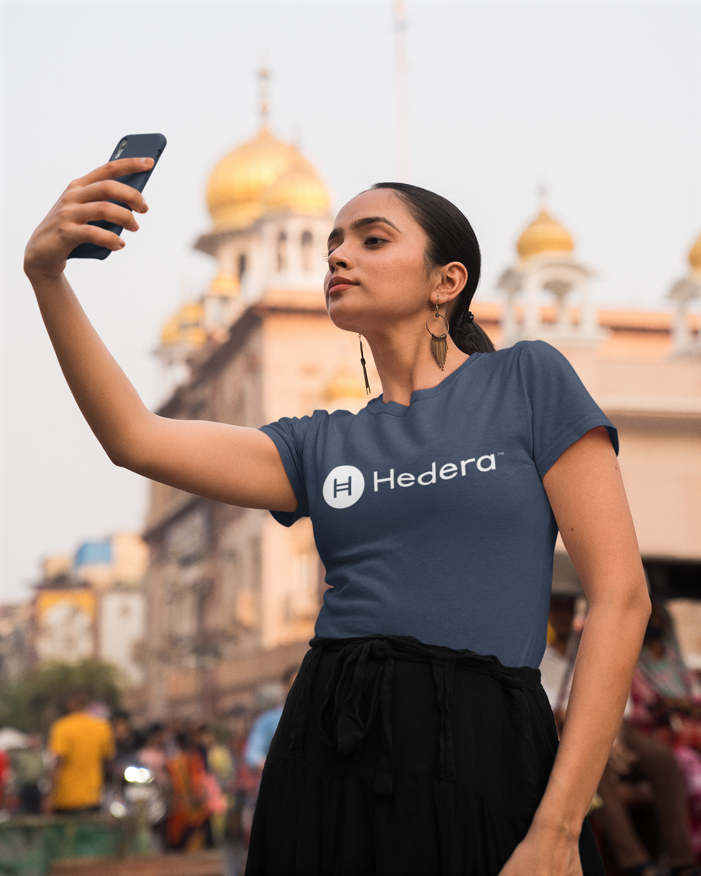 Woman taking a selfie wearing a Hedera t-shirt with a blurred background of a building with domes.